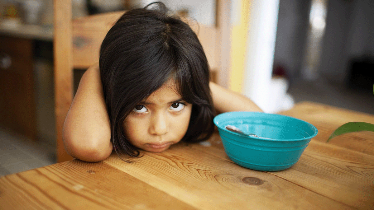 A young child sits at a table with her hands behind her head and a sad expression. An empty bowl sits in front of them on the table.