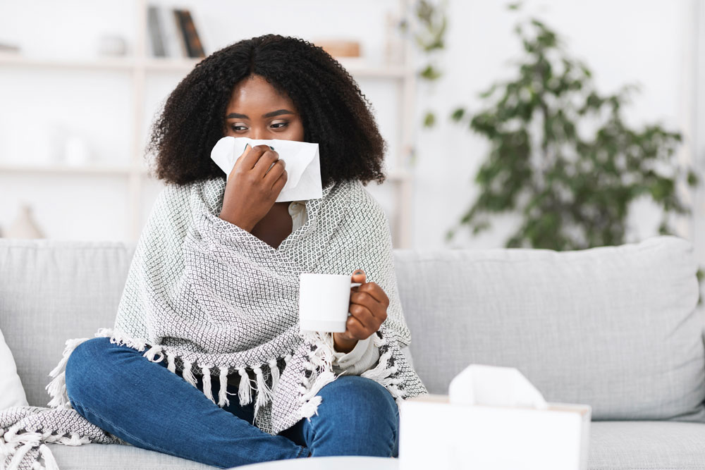 woman sitting on sofa wrapped in blanket blowing nose in tissue