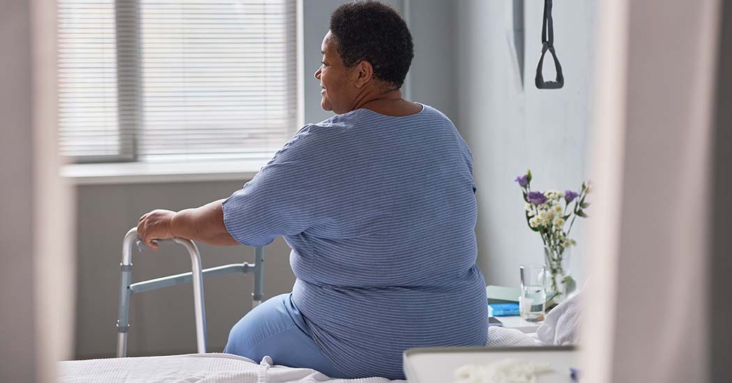 A person of color sits on the edge of a hospital bed. They are holding onto a walker and looking out the window.