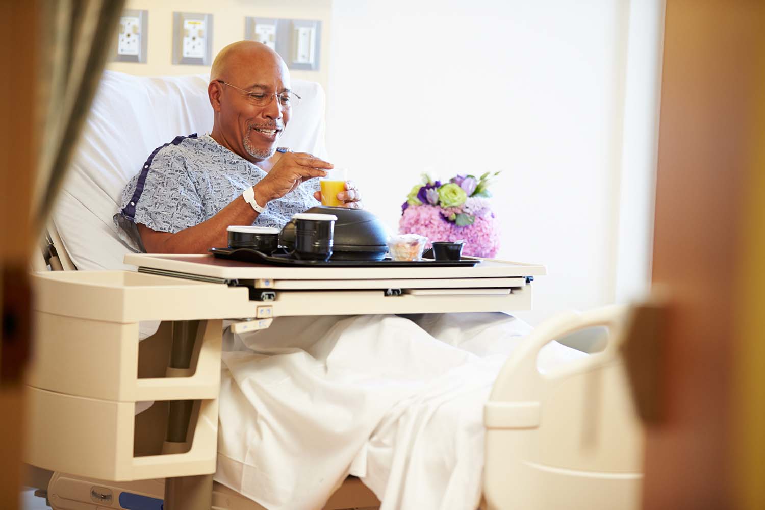 A male patient sits up in a hospital bed as he opens containers of food on a tray table