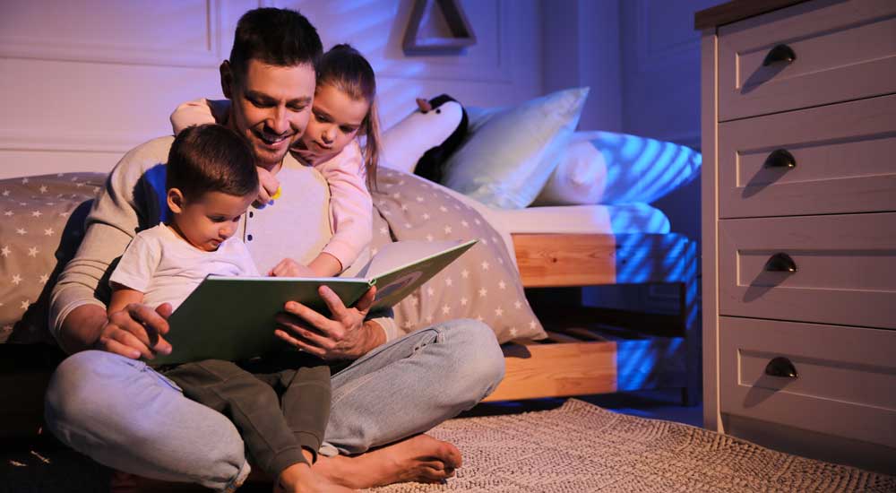 man sitting on floor with two kids reading book