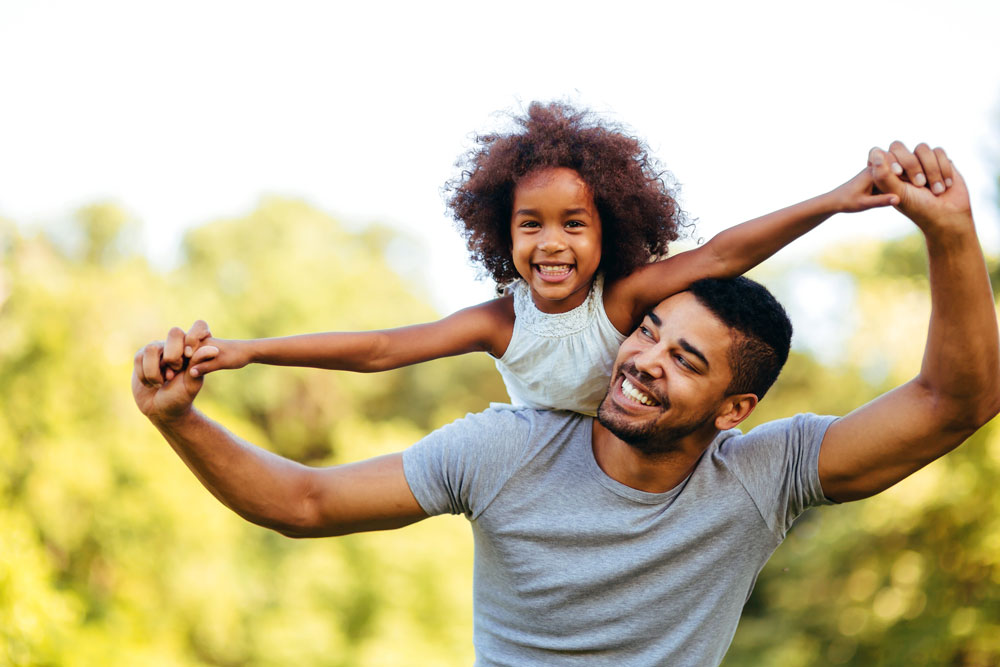 A man smiles as he holds a child up on his shoulders.