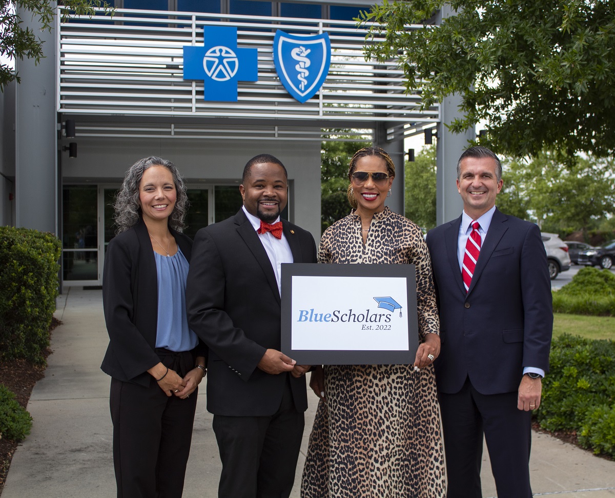 Four people stand in front of a BlueCross building holding a plaque with the BlueScholars logo.