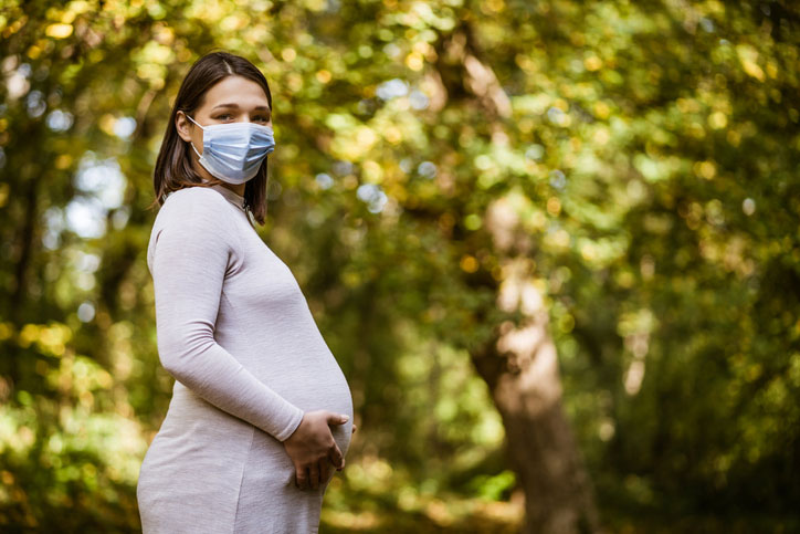 A pregnant woman wearing a face masks poses in front of trees and supports her belly.