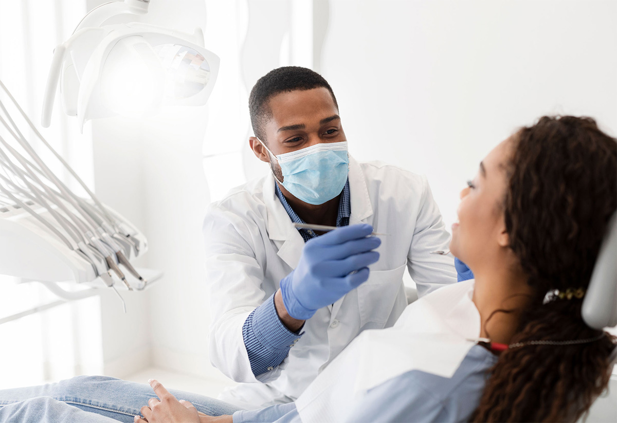 A masked male dentist holds tools as he speaks with a female patient.