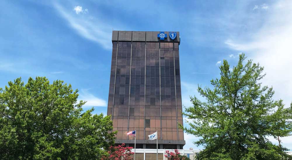 tall building with blue cross logo surrounded by blue sky and green trees
