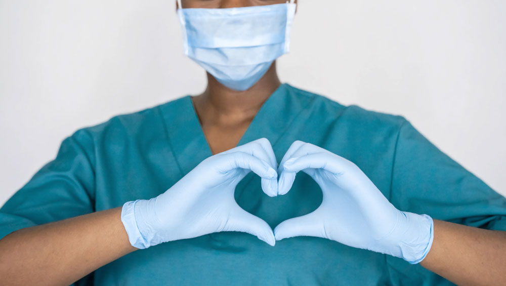 nurse in blue scrubs wearing face mask and gloves makes heart sign with hands