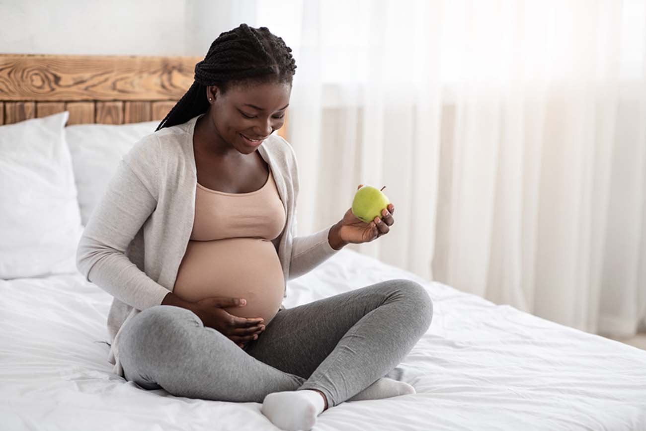 A black pregnant woman sits on a bed and cradles her belly with one hand and holds an apple with the other hand.