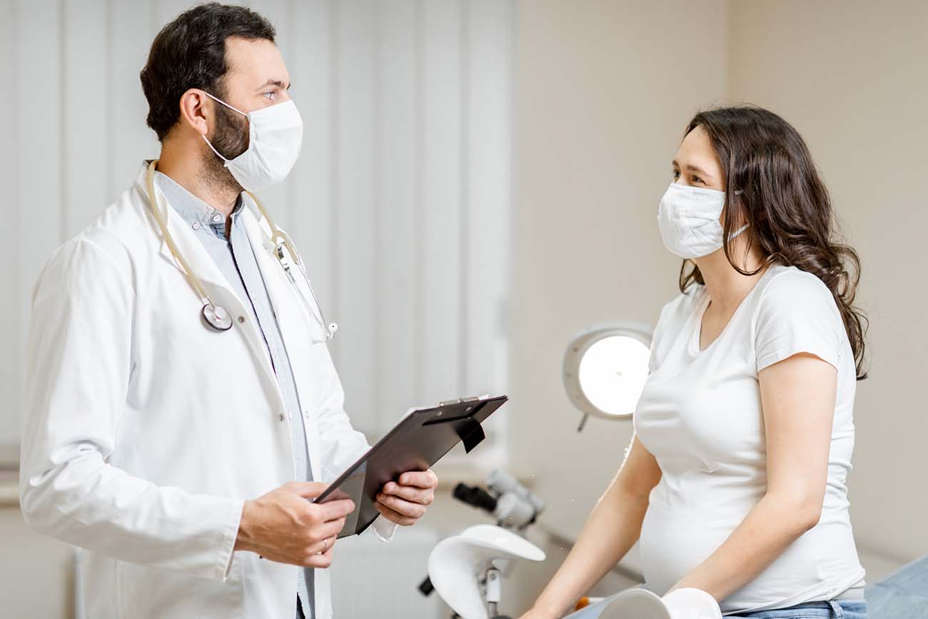 A doctor wearing a face mask and holding a clipboard speaking with a pregnant woman also wearing a mask as she sits on an exam table.