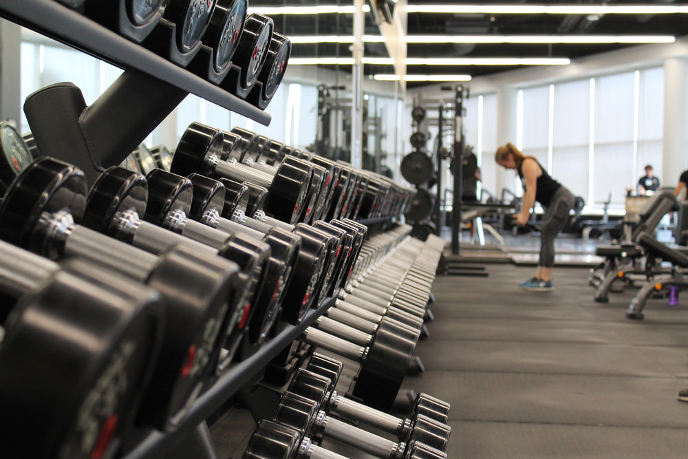 row of dumbbells at gym with woman in distance working out