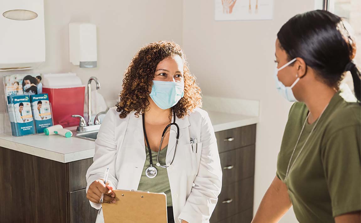 A masked female doctor holds a clipboard and speaks with a masked female patient.