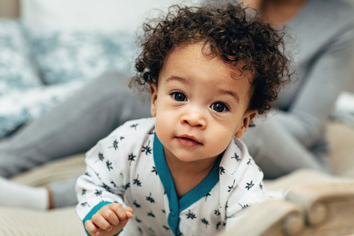 A baby in a white printed onesie looks up into the camera.