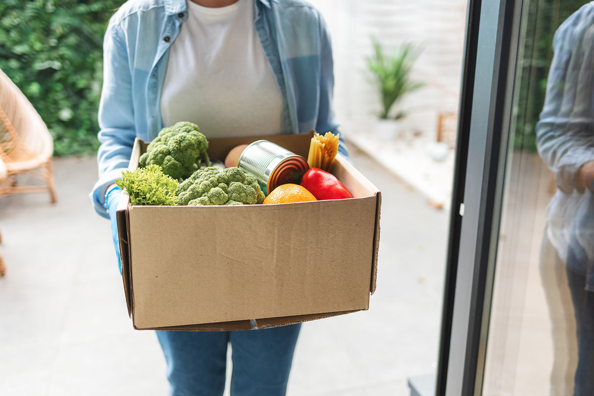 A person carries a cardboard box full of fresh produce and canned goods through a doorway
