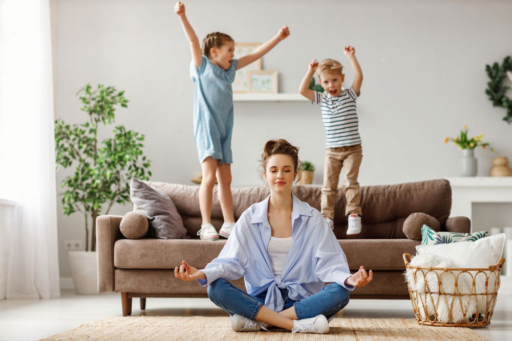 mom sits on floor meditating with kids jumping on sofa